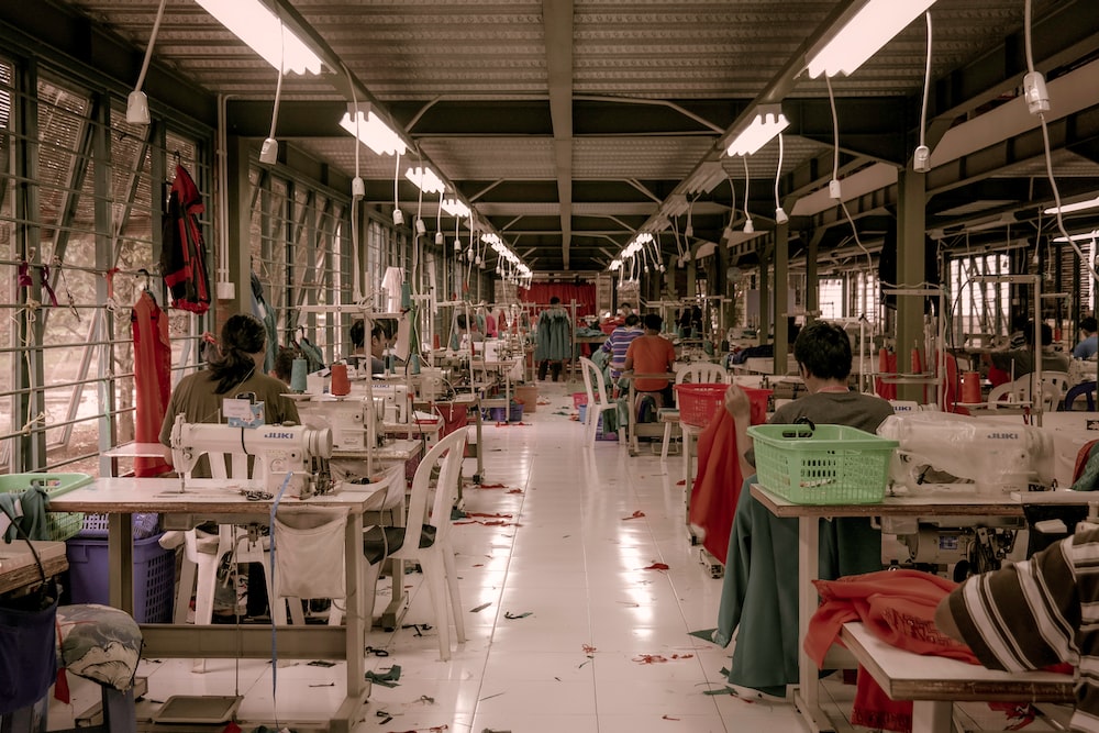 Foto de uma fábrica de roupas. É possível ver várias pessoas, máquinas de costurar e retalhos ao chão.